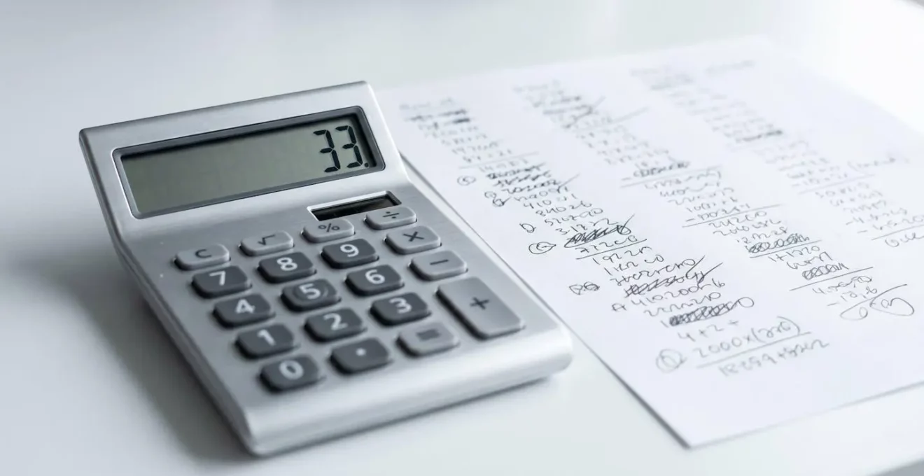 Gros plan sur calculatrice affichant 33 posée sur bureau blanc avec feuille de calculs manuscrits et main tenant stylo